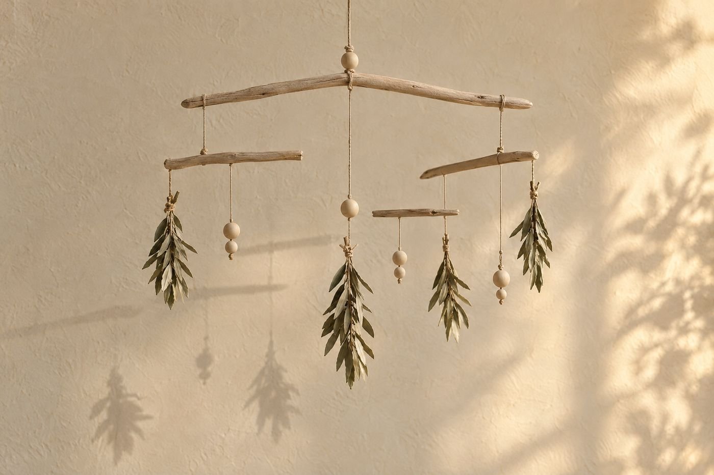 Driftwood mobile hung with dried olive leaves and wooden beads, balanced in front of a cream plaster wall